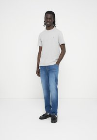 Γκρι κοντομάνικη μπλούζα με στρογγυλή λαιμόκοψη, συνδυασμένη με μπλε τζιν και μαύρα παπούτσια. Η μπλούζα έχει ένα μικρό λογότυπο στο στήθος.