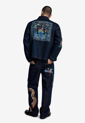 Uomo che indossa una giacca e pantaloni di denim scuro con motivi floreali, animali e di draghi ricamati a colori sulla schiena, sulle maniche e sulle gambe.