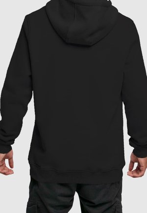 Hombre con sudadera con capucha negra simple y pantalones cargo negros, visto desde la espalda sobre un fondo claro.