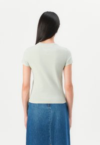 Kvinde med langt mørkt hår iført en lysegrå kortærmet bluse og blå denimnederdel, der står med ansigtet mod en ensfarvet hvid baggrund.