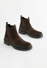 Bocanci Chelsea din suede maro, cu panouri elastice pe laterale, vârfuri rotunjite și tălpi groase din cauciuc negru cu șanțuri adânci.