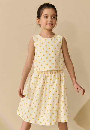 Mladé dievča nosí biele šaty bez rukávov so žltými bodkami a stojí pred jednoliatym béžovým pozadím.