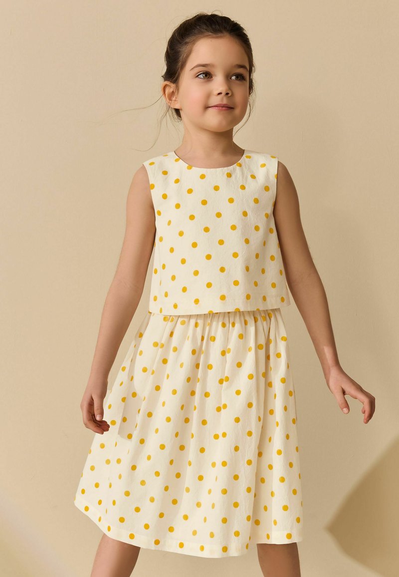 Joven niña con un vestido blanco sin mangas con lunares amarillos, de pie contra un fondo beige liso.