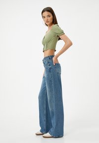 Grünes Crop-Top mit Puffärmeln, kombiniert mit weit geschnittenen blauen Jeans und weißen Turnschuhen, zeigt eine lässige, entspannte Silhouette.