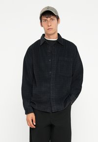 Black checkered button-up shirt in textured fabric, pocket on left chest, worn over a black crew neck t-shirt, paired with dark pants and a cap.