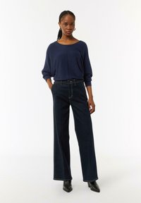 Marineblauer Oversized-Pullover mit langen Ärmeln, kombiniert mit dunkelblauen, weit geschnittenen Jeans. Beide Kleidungsstücke weisen einen lässigen Schnitt und ein minimalistisches Design auf.