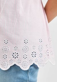 Vaaleanpunainen ja valkoinen raidoitettu paita, jossa on tikattu silmäkansia helma, joka esittää kukkakuvioita. Alhaalla on aallotettu reuna, joka täydentää vaalean denim-kankaan.