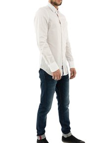 Camisa blanca de manga larga, botón en la parte delantera, con corte ajustado y cuello estándar, combinada con jeans oscuros y zapatillas negras con detalles en claro.