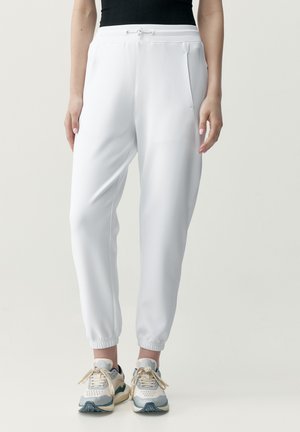 Weiße Jogginghosen mit schmal zulaufenden Beinen und elastischen Bündchen, getragen mit mehrfarbigen weißen und grauen Sneakers und einem schwarzen ärmellosen Oberteil