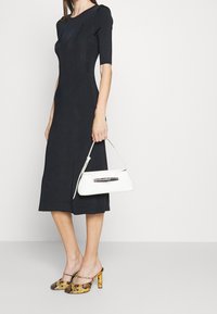 Robe midi noire à côtes avec des manches courtes, associée à un sac à main blanc avec un accent horizontal et des chaussures à talons jaunes à motifs.