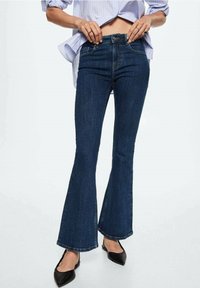 Jeans de mezclilla oscuros de corte acampanado con una cintura alta y detalles de costura mínimos, combinados con tacones negros de punta.
