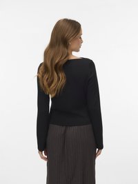 Žena s dlouhými vlnitými světle hnědými vlasy, oblečená v černém tričku s dlouhým rukávem a hnědých kalhotách s proužky, otočená zády na bílém pozadí.