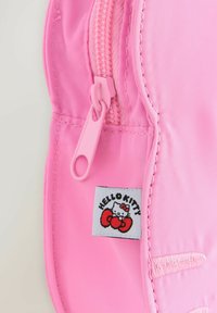 Tissu rose avec fermeture éclair et étiquette Hello Kitty cousue, présentant la tête du personnage et un nœud rouge.