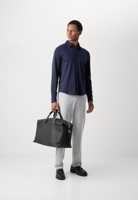 Borsa da viaggio in pelle nera con superficie testurizzata, accessori in argento e manici doppi. Il modello indossa una camicia blu con bottoni e pantaloni grigi chiari.