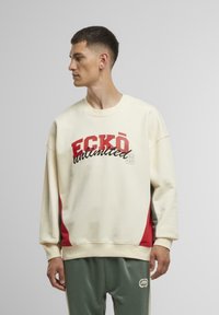 Beige Sweatshirt mit roten Akzenten, versehen mit auffälligem Drucktext. Das Design hat eine lockere Passform und gerippte Bündchen. Kombiniert mit grünen Jogginghosen.