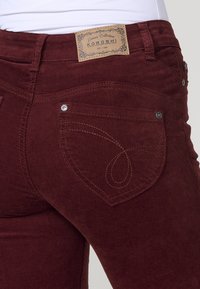 Close-up van bordeauxkleurige corduroy broek met achterzak, versierd met stiksel en een tan leren merkpatch op de tailleband.