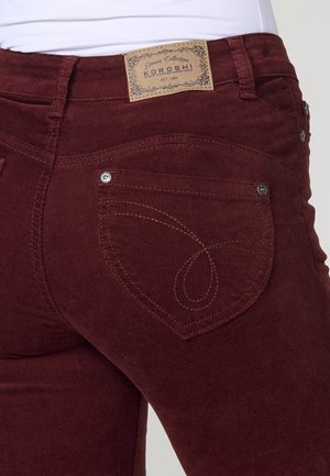 Nahaufnahme von kastanienbraunen Cordhosen mit Gesäßtasche, die dekorative Stickerei aufweist, und einem hellbraunen Leder-Brandpatch am Bund.