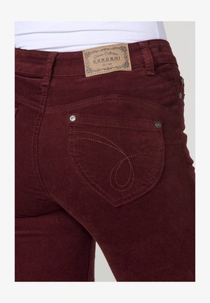 Lähedal pilt tumepunast kardinakanga pükstest, mille tagaküljel on dekoratiivne õmblus ja talje peal pruun nahkbrändi silt.