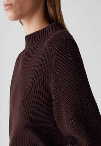 Pull en tricot marron foncé, doté d'un col montant, d'un ribbing texturé et d'une coupe ample avec des coutures diagonales sur les manches.