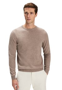 Brun stickad tröja med rundhals, ribbade ärmslut och nederkant. Bärs över ljusa byxor med en mjuk och texturerad yta.