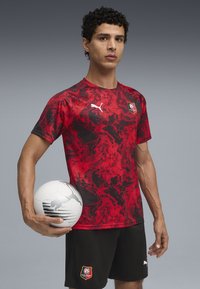 Punase ja musta mustriga spordipoliis, millel on lühikesed varrukad, koos mustade lühikesed pükstega, millel on logo; ühes käes hoitakse valget jalgpalli.