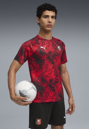 Rød og sort mønstret sports skjorte med korte ærmer, sammen med sorte shorts med et logo; en hvid fodbold holdes i den ene hånd.