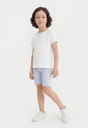 Παιδί με σγουρά μαλλιά που φοράει λευκό μπλουζάκι, ανοιχτό μπλε σορτς και λευκά αθλητικά παπούτσια, στέκεται στο ένα πόδι μπροστά σε απλό φόντο.