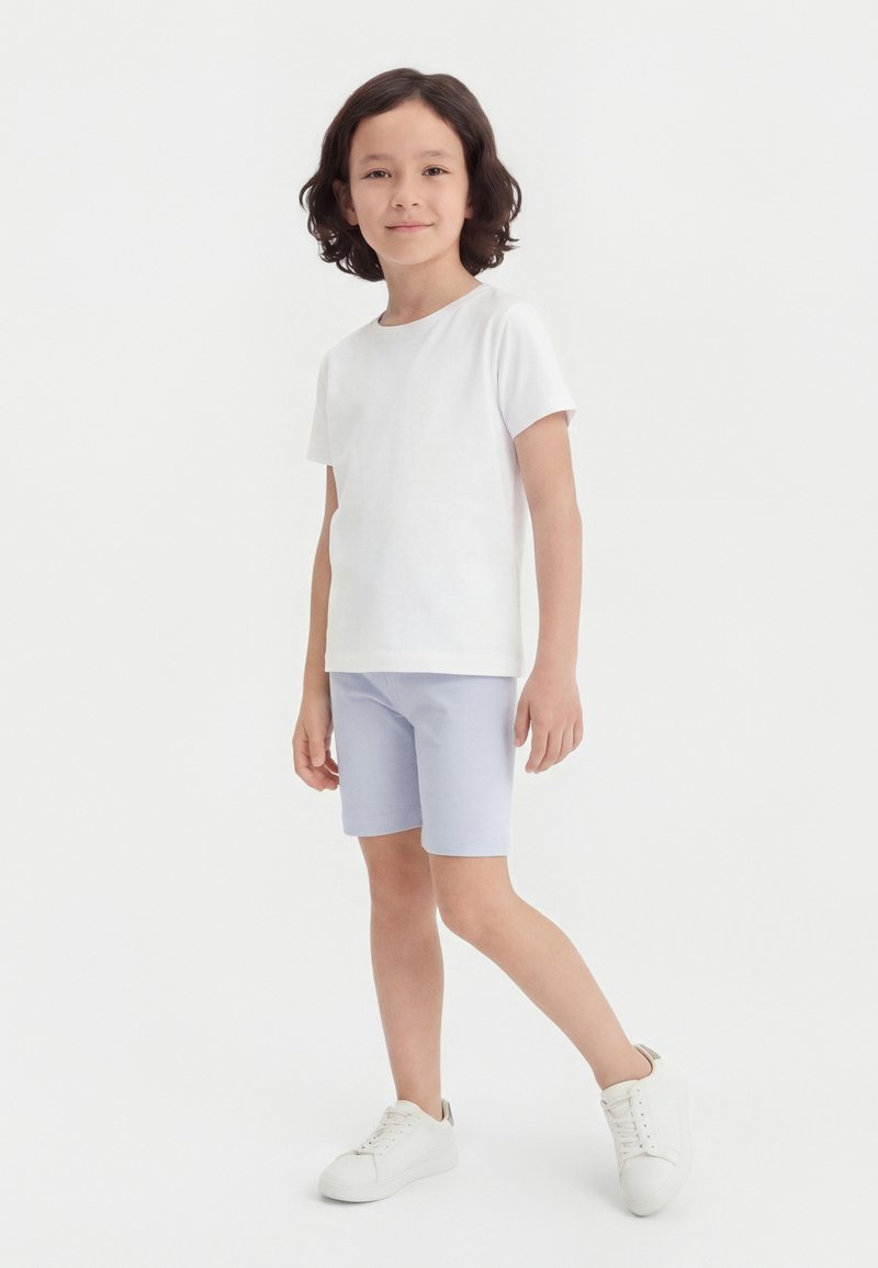 Παιδί με σγουρά μαλλιά που φοράει λευκό μπλουζάκι, ανοιχτό μπλε σορτς και λευκά αθλητικά παπούτσια, στέκεται στο ένα πόδι μπροστά σε απλό φόντο.