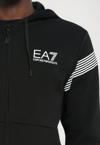 Juodas zip-up pūkinė striukė, pagaminta iš minkštos medžiagos, su logotipu krūtinėje ir baltomis horizontaliomis juostomis ant pečių. Reguliuojama kapota.