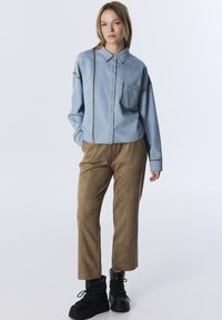 Lichtblauwdenimshirt met contrasterende bruine stiksels, knoopsluiting, één borstzak; gecombineerd met tan suède broek en zwarte laarzen.