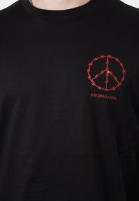 Maglietta nera con un simbolo della pace rosso fatto di filo spinato e la parola "PROPAGANDA" stampata sotto sul lato sinistro del petto.