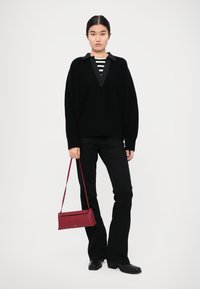 Black knitted sweater with a V-neck and ribbed texture, layered over a striped shirt. Black flared pants; holding a red leather handbag.