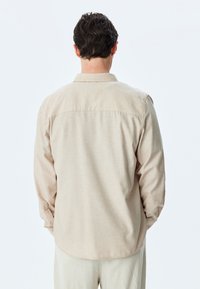 Camisa de manga larga beige con yugo en la parte posterior y dobladillo redondeado. Elaborada con una tela texturizada, cuenta con puños abotonados y un cuello clásico.