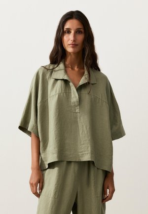 Frau mit langen dunklen Haaren, die ein lockeres olivgrünes Leinenoberteil mit Kragen und passende Hose trägt, steht vor einem schlichten hellen Hintergrund.