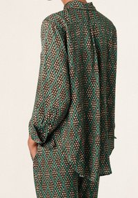Blusa a maniche lunghe con motivo a diamante in marrone, teal e bianco, dotata di colletto, piega sul retro e vestibilità rilassata. Tessuto morbido con caduta.