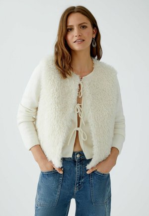 Witte nepbontjas met een ronde hals en striksluiting aan de voorkant, gedragen over een crèmekleurige trui; gecombineerd met blauwe jeans.