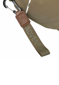 Détail d'un sac en tissu beige avec une pièce en cuir et une sangle en toile verte portant le mot "AVENTURES" en lettres majuscules. Mousqueton métallique attaché.