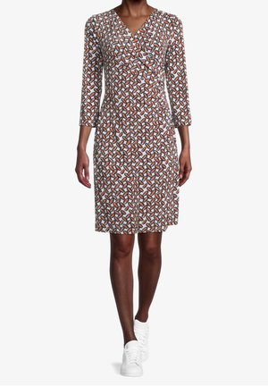 Kvinde iført knælængde omslagskjole med lange ærmer og geometrisk mønster i orange, sort og hvid, kombineret med hvide sneakers.