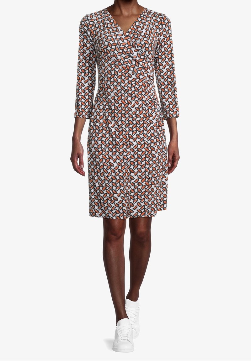 Femme portant une robe portefeuille à manches longues, longueur genou, avec un motif géométrique orange, noir et blanc, associée à des baskets blanches.