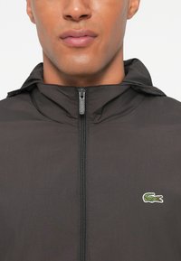 Giacca leggera nera con cappuccio, chiusura con zip frontale completa e logo Lacoste ricamato sul petto sinistro in verde. Texture liscia.