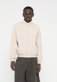 Ελαφριά μπεζ μπέρμπον μπουφάν από λείο ύφασμα, με ψηλό γιακά, φερμουάρ μπροστά, ελαστικά μανίκια και ζώνη στη μέση. Κατάλληλο για στρώσεις.