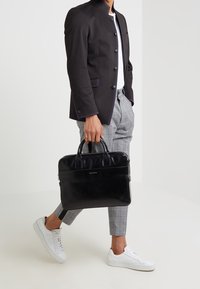 Black leather briefcase with dual handles, smooth texture, zip closure, and a front logo. Pairing with grey plaid trousers and white sneakers.