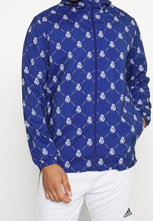 Niebieska kurtka na zamek z białymi powtarzającymi się logo Realu Madryt, noszona przez osobę w białych spodniach Adidas z widocznym logo Adidas.