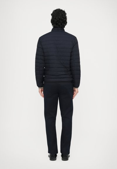 Temno modra, žametno obložena puhasta jakna z visokim ovratnikom, nošena čez črne hlače. Gladek material z navpičnimi šivi, ki poudarjajo oprijeto obliko.