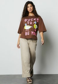 Tricou maro supradimensionat cu grafică colorată cu rațe și textul „DUCKS TO BE YOU.” Asortat cu pantaloni bej și adidași multicolori.