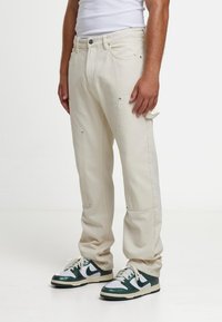 Beežid puuvillased cargo-püksid küljetaskute, metallist detailide, lõdva lõike ja kergelt kitsenevate säärtega. Kanditud roheliste ja valgete tennistega.