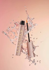 Tubo di mascara rosa oro con wand nera, parzialmente immerso in acqua che schizza su uno sfondo rosa chiaro. Scritta in oro visibile.