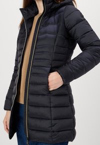 Veste doudoune noire isolée avec capuche, présentant une texture matelassée, une fermeture éclair frontale intégrale et des poches latérales. Tissu doux, arrive au milieu de la cuisse.