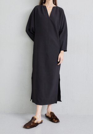 Kobieta w długiej czarnej sukience z bocznymi rozcięciami i brązowymi futrzanymi wsuwanymi butami, stojąca na tle jednolitej jasnoszarej ściany.