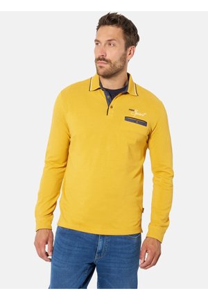 Mann trägt ein gelbes langärmeliges Poloshirt mit marineblauen Abschlüssen und "Free Spirit"-Text, kombiniert mit blauen Jeans, steht vor einem schlichten Hintergrund.